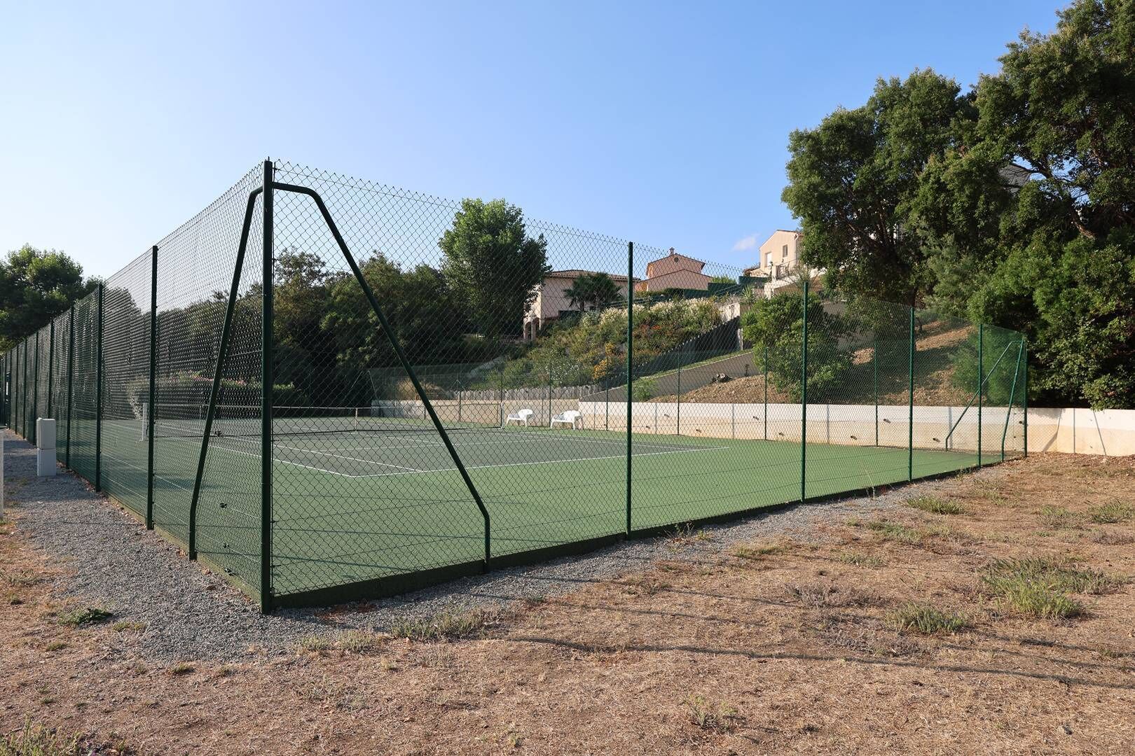 A tennis court enclosed by a chain-link fence with a gravel path in front.