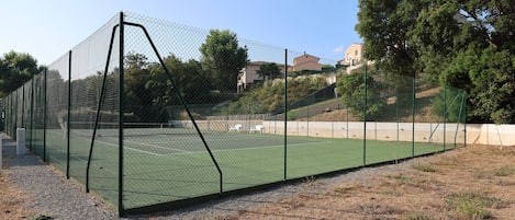 A tennis court enclosed by a chain-link fence with a gravel path in front.