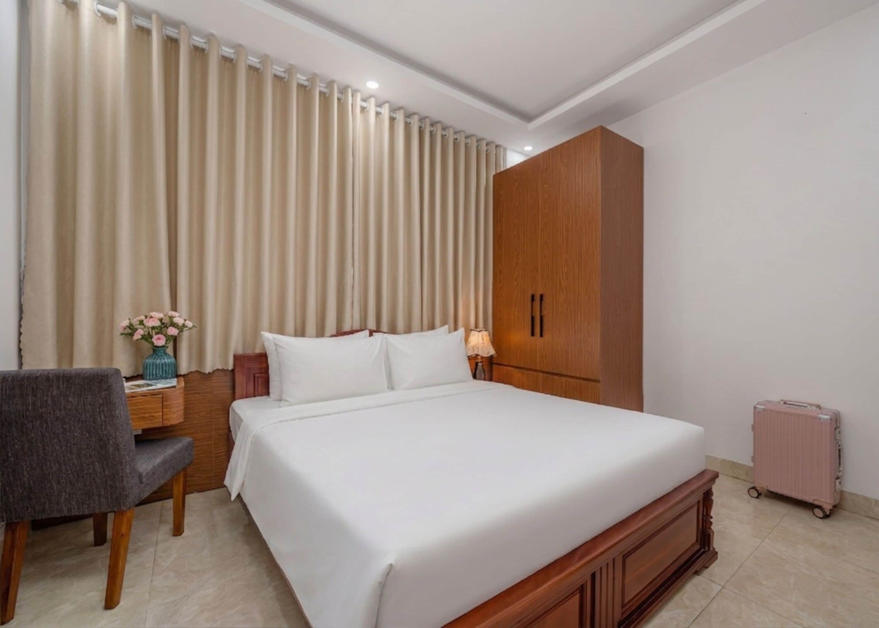 A hotel room with a large bed, a wooden wardrobe, a desk with a chair, and a suitcase.