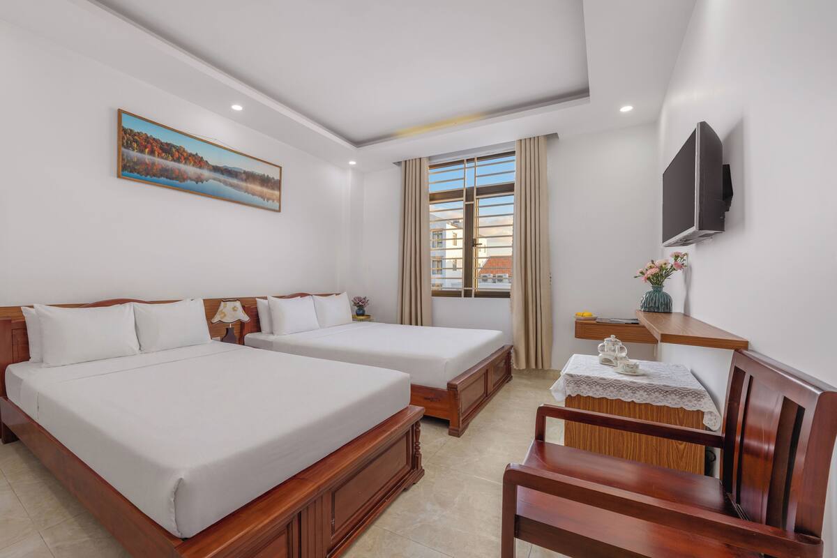 Quarto de hotel com duas camas, uma televisão, uma janela com cortinas, uma mesinha com um vaso e uma cadeira.