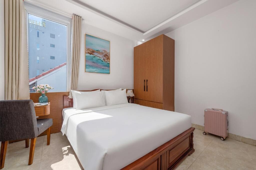 A hotel room with a bed, a desk with a chair, a wardrobe, a window with curtains, and a painting on the wall.