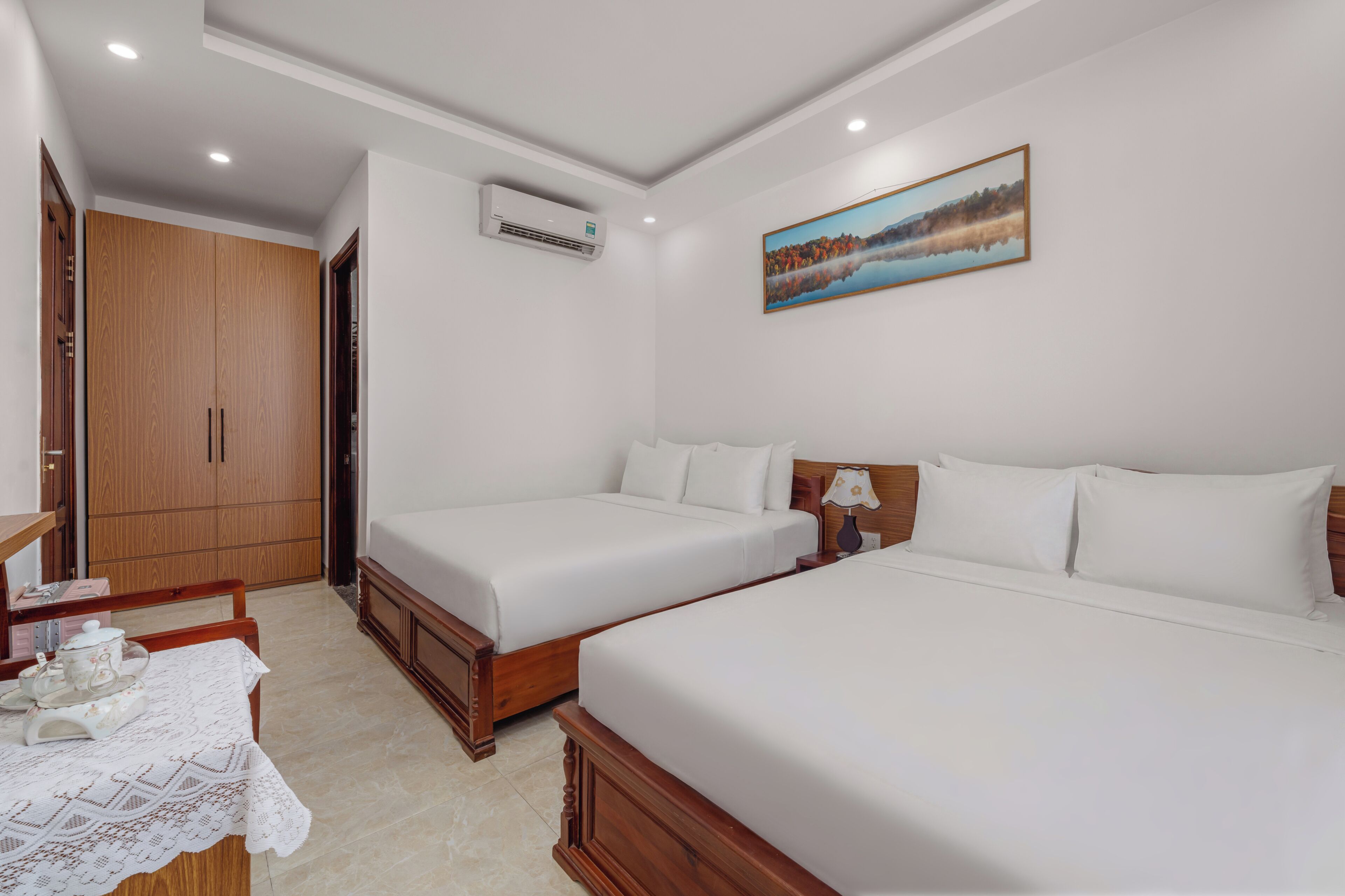 A hotel room with two beds, a wooden wardrobe, a painting on the wall, and a table with a lace tablecloth.