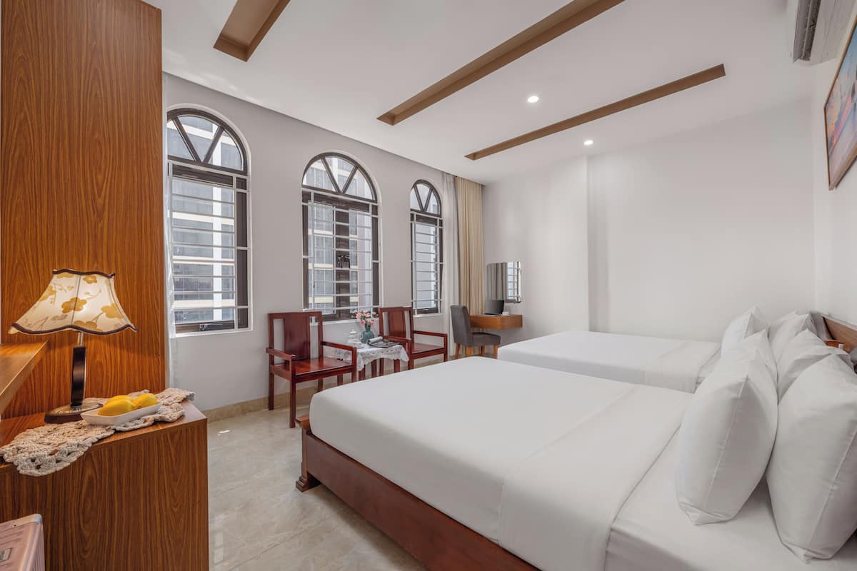 Quarto de hotel com duas camas, uma mesa com abajur, uma cadeira e uma pequena mesa com um vaso.