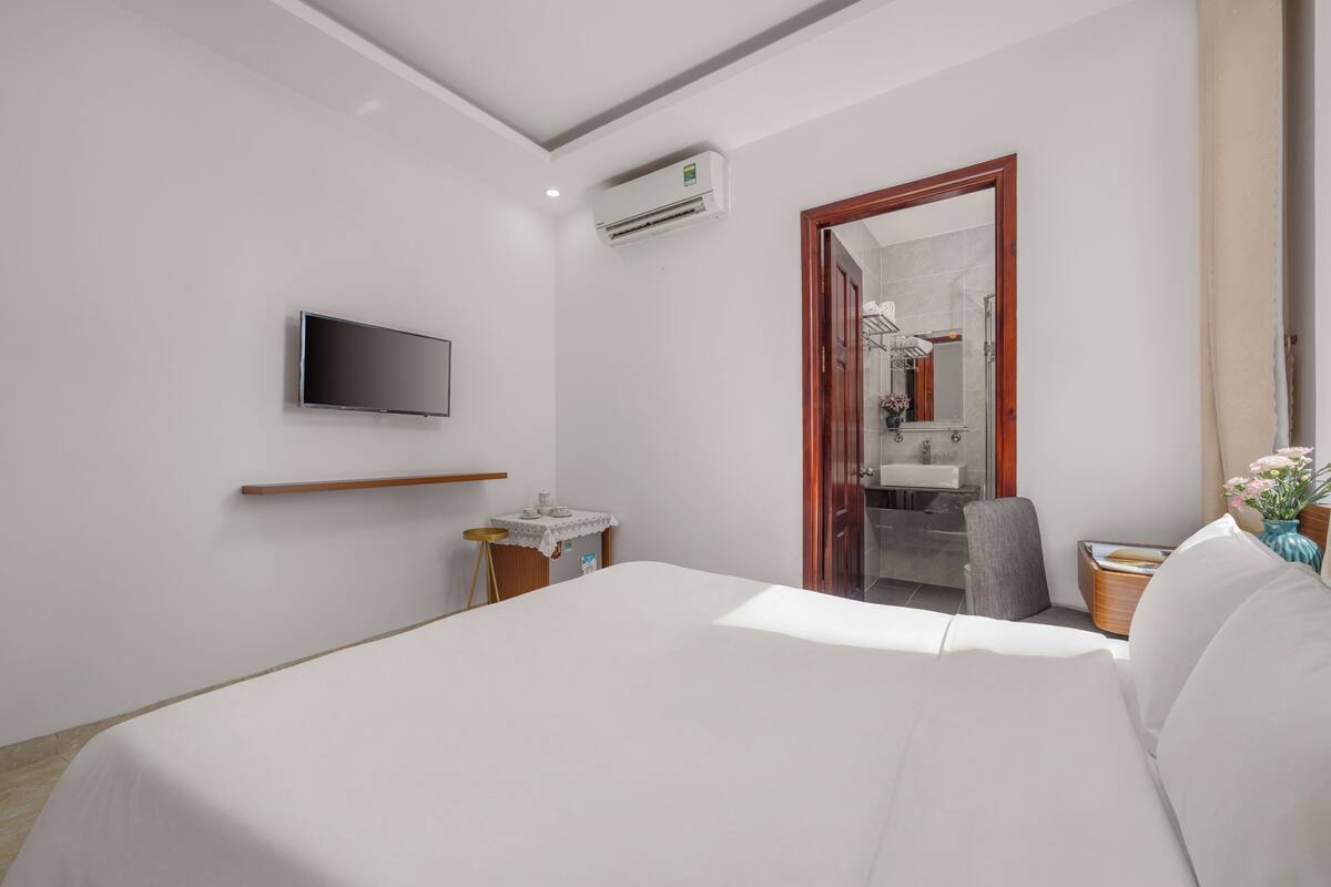 Quarto de hotel com cama, TV fixada na parede, uma mesinha com abajur e banheiro visível através de uma porta aberta.