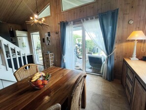 Ein Holzinnenraum mit Esstisch, Korbstühlen, einer Obstschale, einer Küchenzeile und einer Glasschiebetür, die auf eine Terrasse im Freien führt.