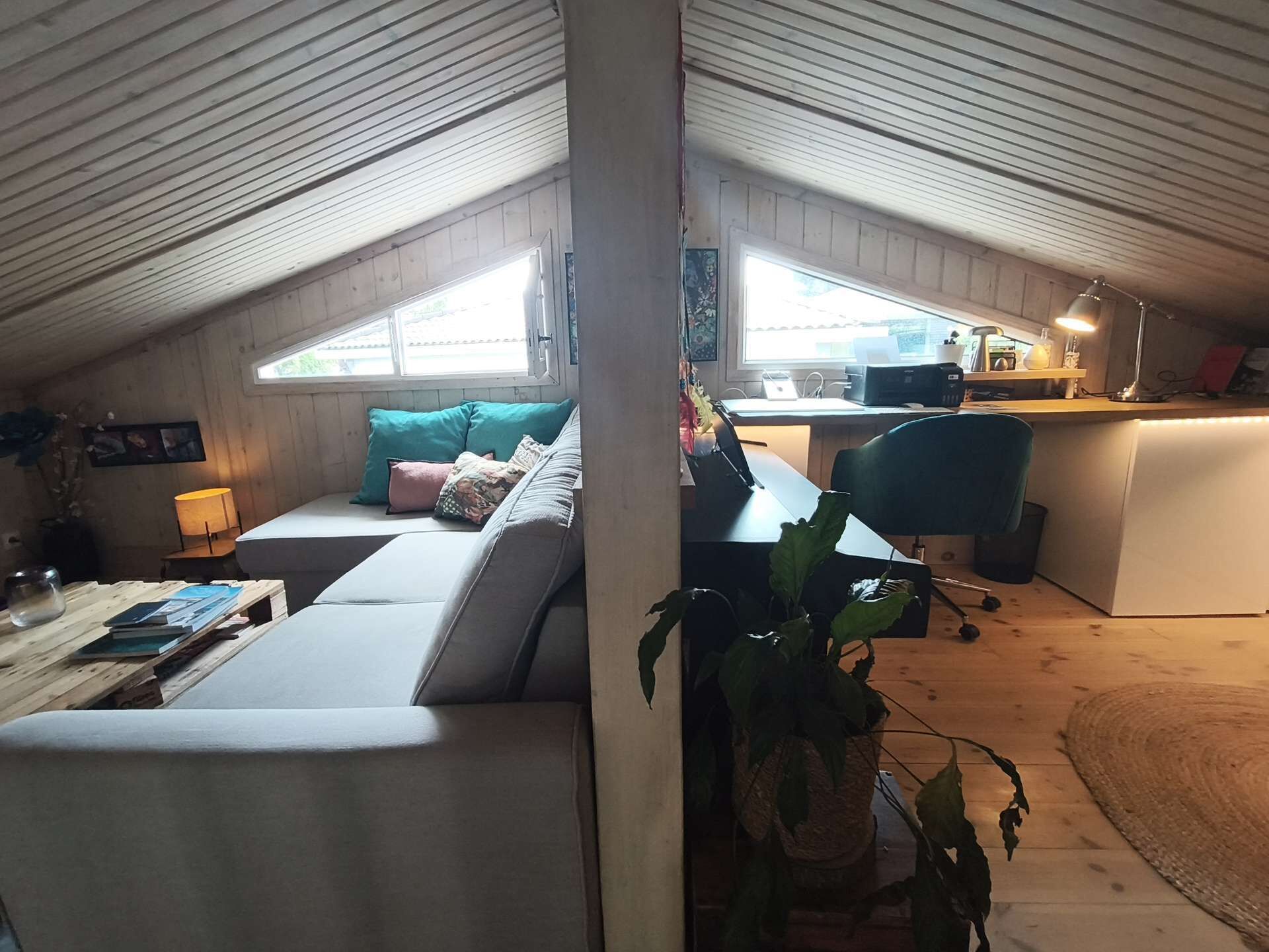 A loft-style room with a high ceiling, a sofa with cushions, a desk with a chair, and a potted plant.
