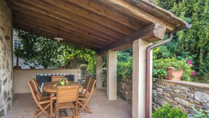 Eine überdachte Terrasse mit Holztisch und -stühlen, Steinwänden und Kübelpflanzen.