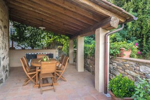 Eine überdachte Terrasse mit Holztisch und -stühlen, Steinwänden und Kübelpflanzen.