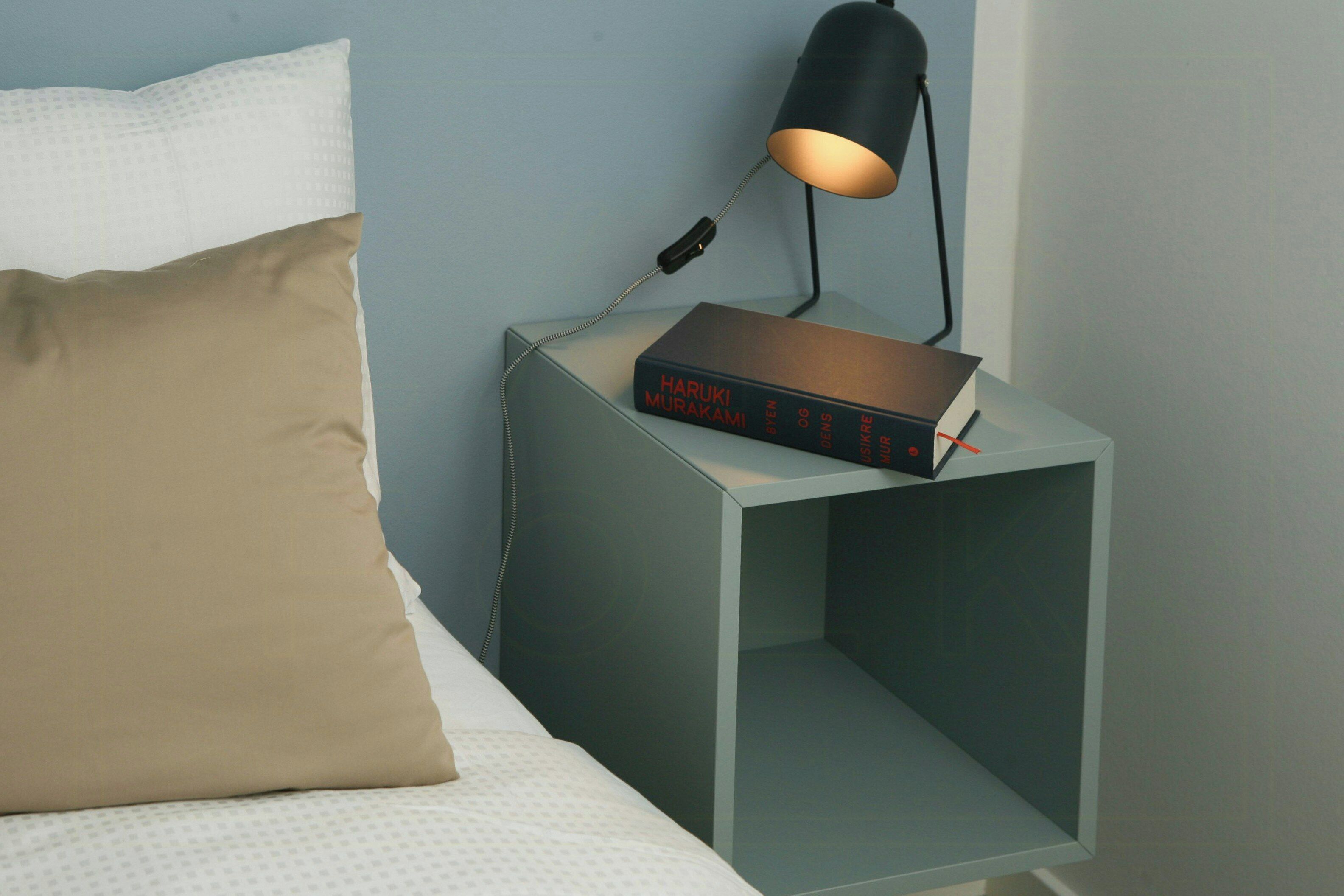 Un lit avec un oreiller blanc, un oreiller couleur fauve et une table de chevet sur laquelle se trouve un livre intitulé « HARUKI MURAKAMI » et une lampe.