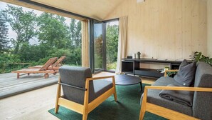 En modern inredning med ett träpanelat tak, stora fönster, en soffa, en fåtölj och en uteplats med solstolar.