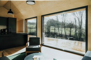 Ein modernes Interieur mit Küche, Sitzgelegenheit und großen Fenstern, die einen Blick auf eine Holzterrasse und Bäume bieten.
