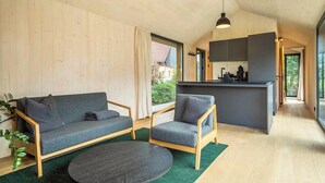 En modern inredning med en soffa, fåtölj och ett runt soffbord i ett ljust rum med träväggar och ett köksutrymme.