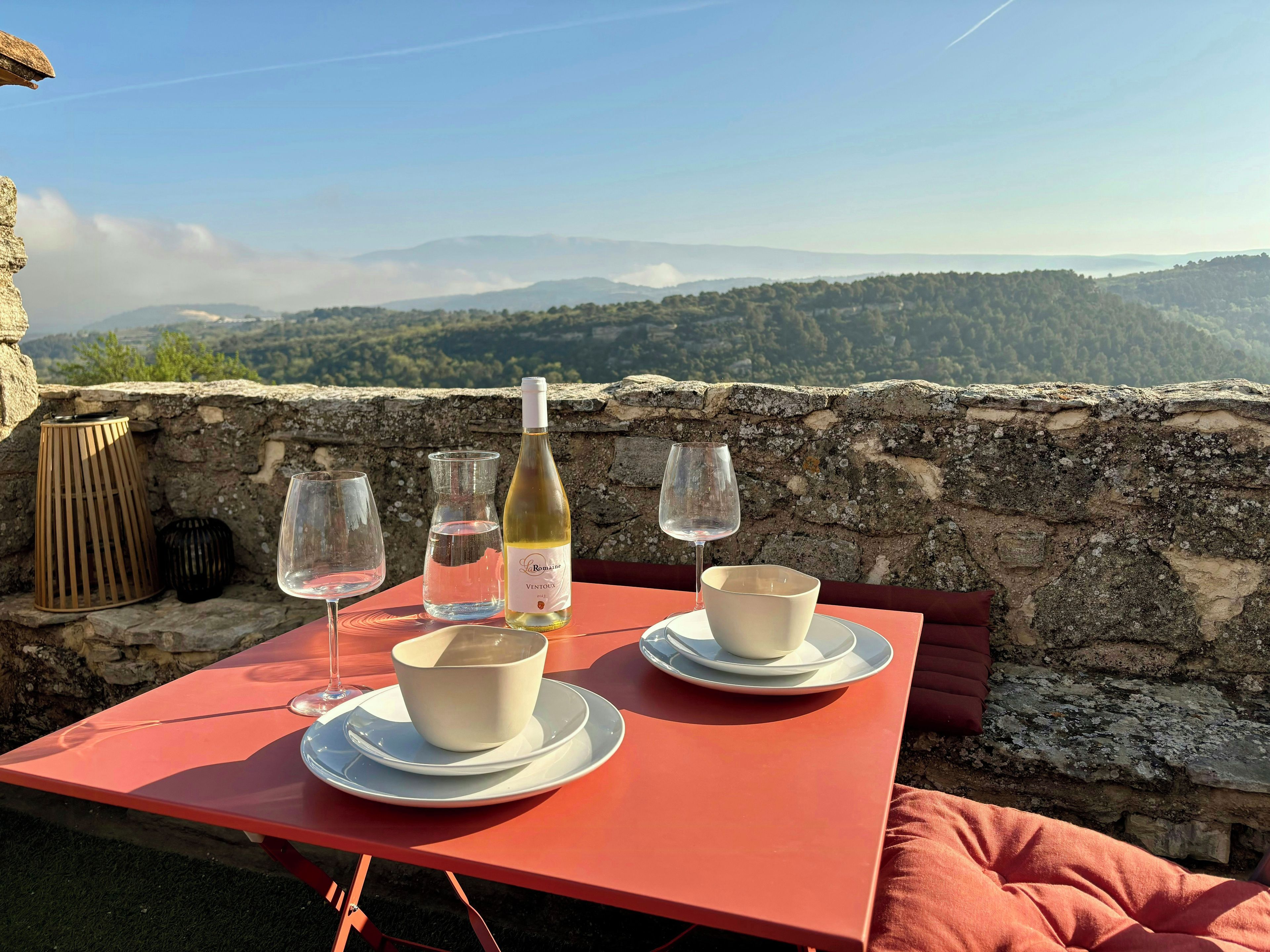 A table with a bottle of wine, glasses, and a bowl set against a scenic mountain backdrop.