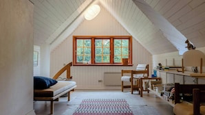 Ein gemütliches Dachzimmer mit geneigter Decke, einem großen Fenster, einem Bett, einem Schreibtisch und Regalen mit Büchern und persönlichen Gegenständen.