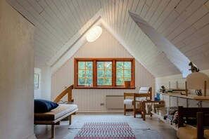Ein gemütliches Dachzimmer mit geneigter Decke, einem großen Fenster, einem Bett, einem Schreibtisch und Regalen mit Büchern und persönlichen Gegenständen.