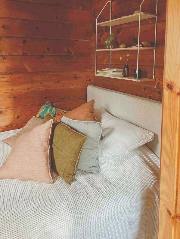 A wooden cabin room with a bed, pillows, a shelf with books and decorative items. - Holiday home with private garden in Ugglehult (Hjärnarp)