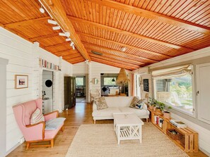A cozy room with a wooden ceiling, a white sofa, a pink chair, a small white table, and a window with a view of greenery. - Holiday home with private garden in Ugglehult (Hjärnarp)