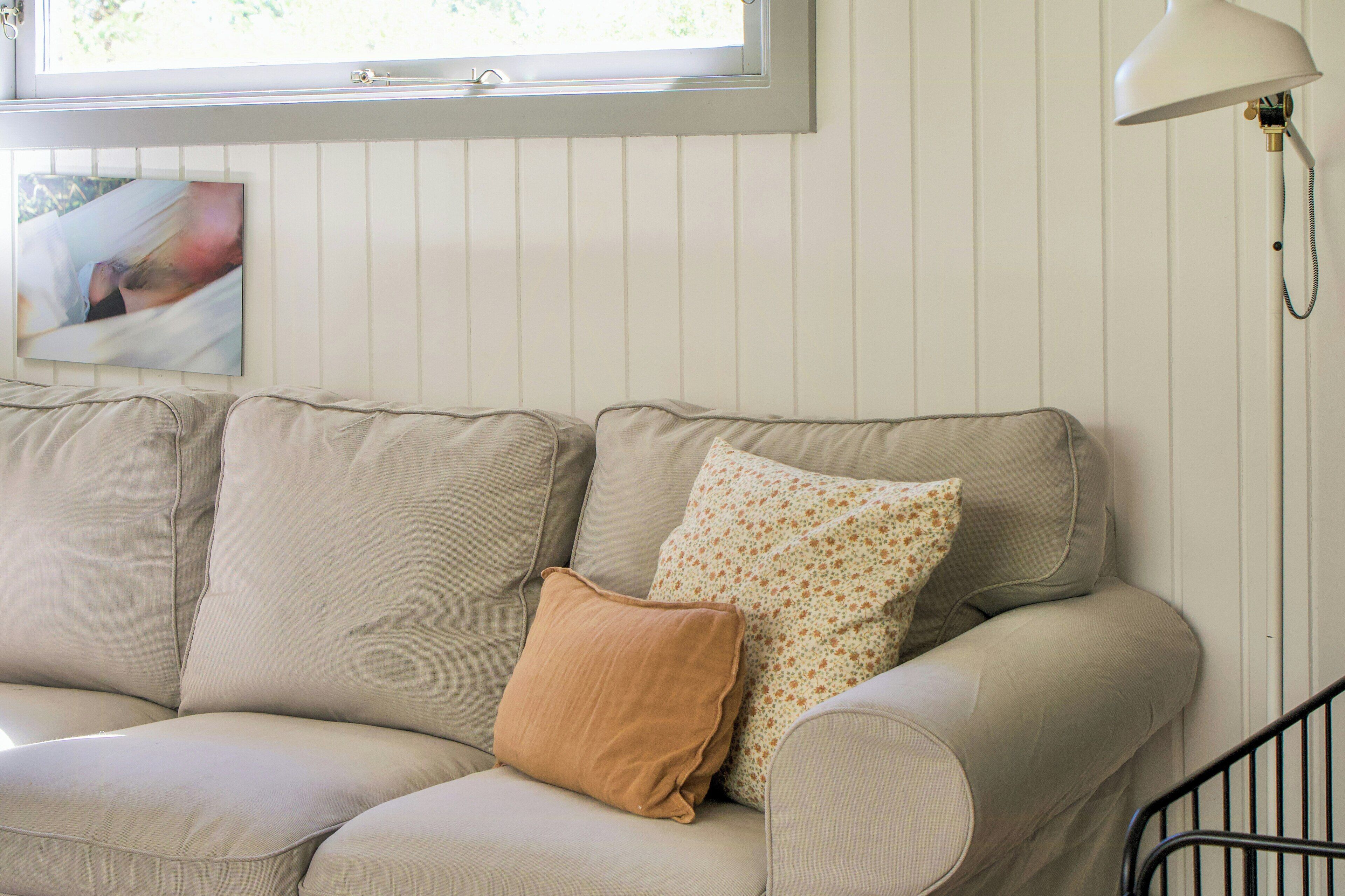 A beige sofa with a brown pillow and a floral pillow, a white lamp, and a framed picture on the wall.