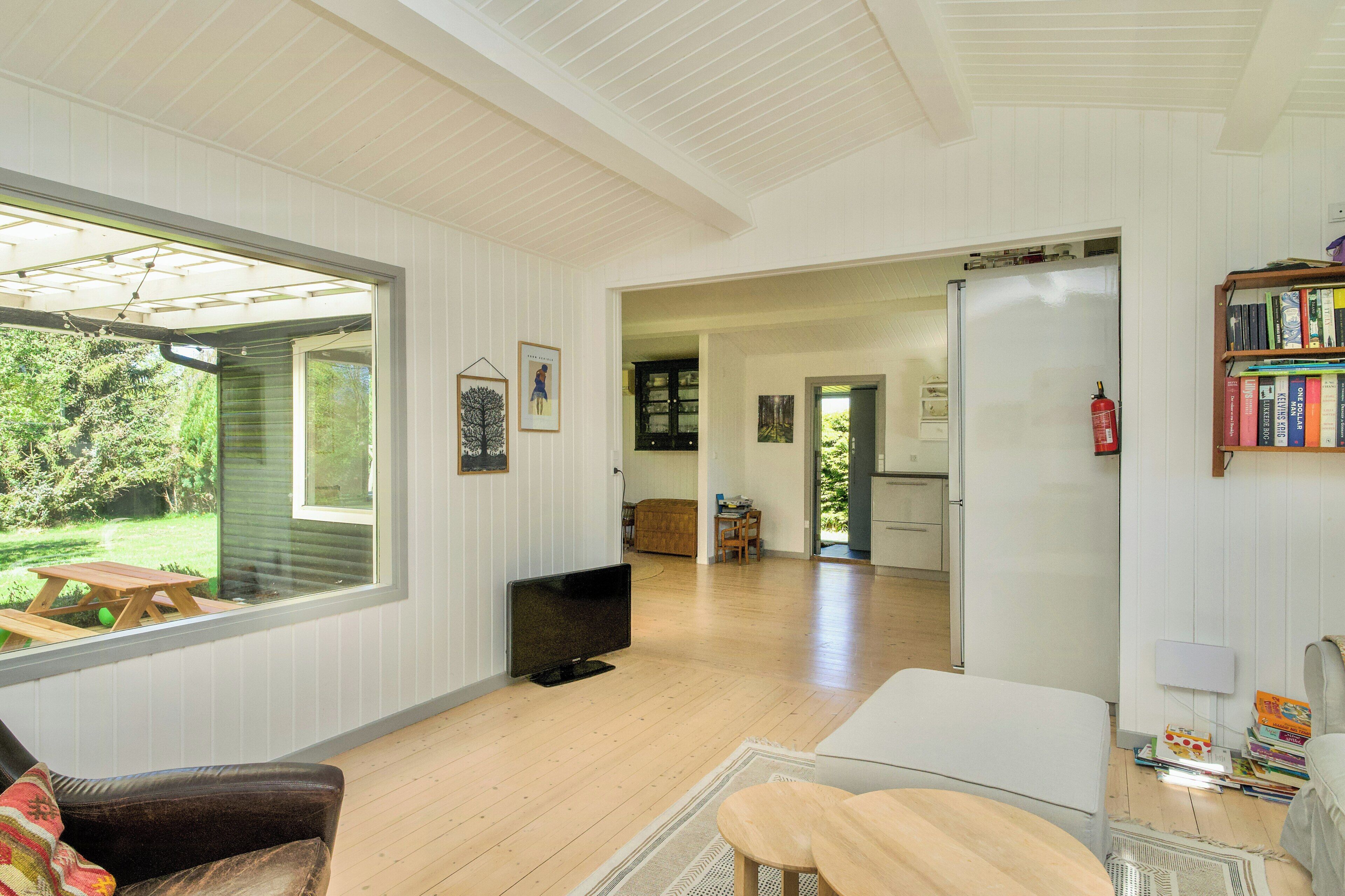 A spacious living room with a large window, a television, a wooden dining table, and a bookshelf.