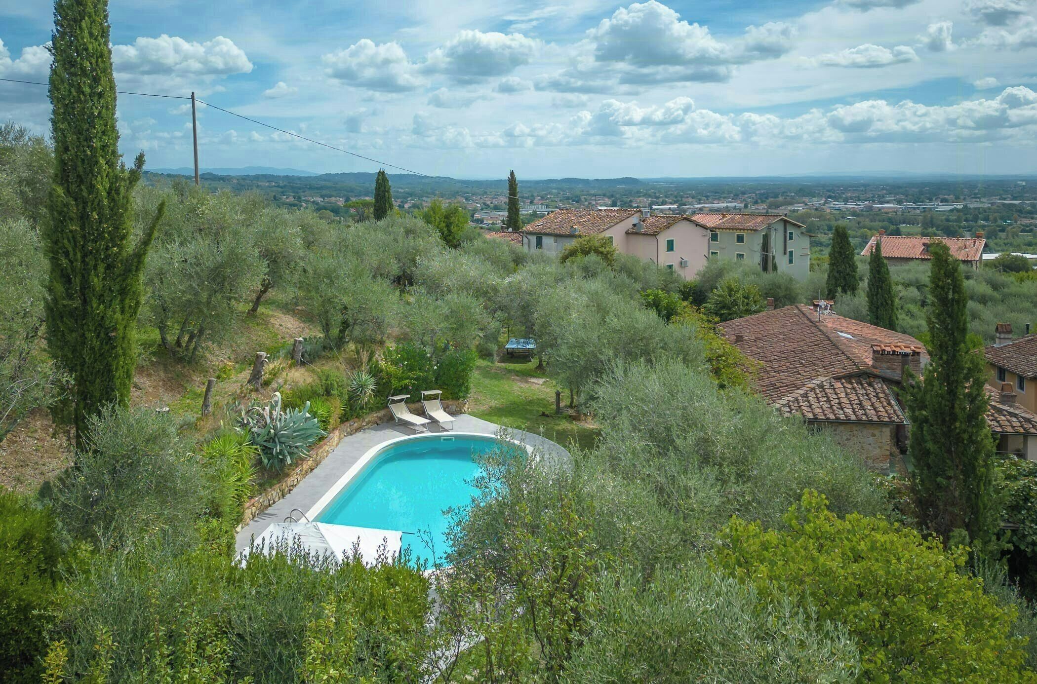 A residential area with a swimming pool surrounded by greenery, houses with red-tiled roofs, and a distant view of a town.