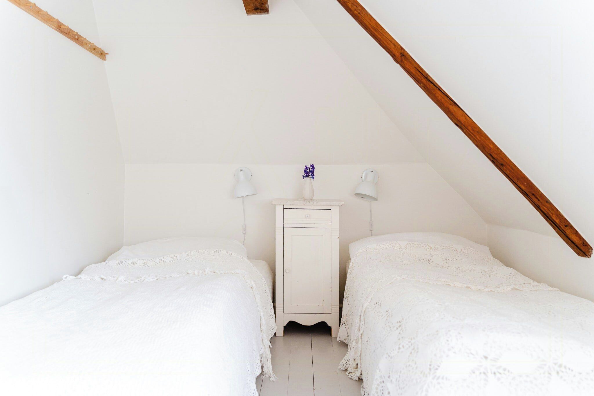 A small, white, attic-style bedroom with two beds, a nightstand, and a wooden beam ceiling.