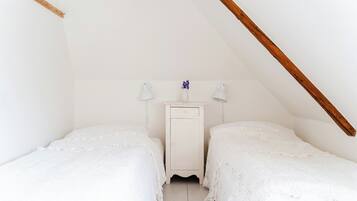 A small, white, attic-style bedroom with two beds, a nightstand, and a wooden beam ceiling.