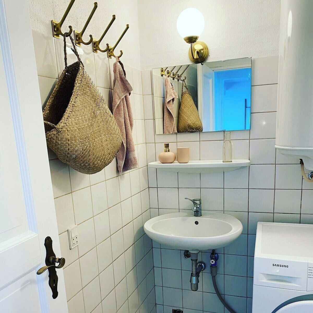 Un baño con lavamanos blanco, un espejo, un perchero con bolsos y una lavadora Samsung.