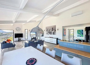 A modern living room with a dining area, a kitchen, and a large window offering a city view. - Modern villa with sea views and infinity pool (Les Adrets-de-l'Estérel)