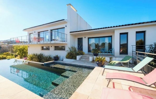 A modern two-story house with a swimming pool, outdoor seating, and a view of the surrounding landscape. - Modern villa with sea views and infinity pool (Les Adrets-de-l'Estérel)