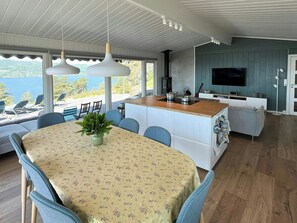 Eine moderne Küche mit Kochinsel, Essbereich mit Stühlen, einem an der Wand montierten Fernseher und Blick ins Freie.