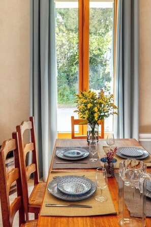 Une table à manger dressée pour quatre personnes, avec des assiettes, des couverts et des verres, un vase rempli de fleurs jaunes et une vue sur la verdure par la fenêtre.