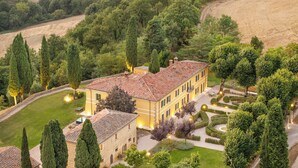 A large, yellow building with a tiled roof surrounded by well-maintained gardens and tall cypress trees.