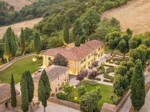 A large, yellow building with a tiled roof surrounded by well-maintained gardens and tall cypress trees.