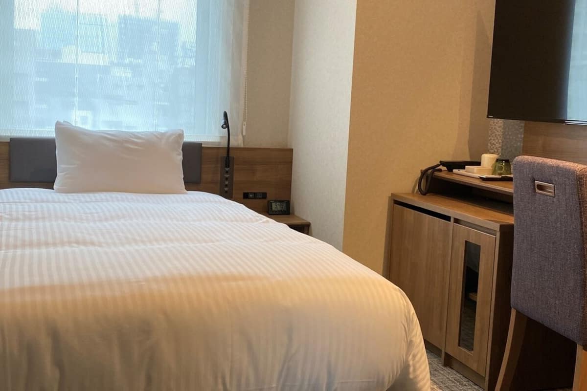 A hotel room with a bed, a desk with a chair, a television, and a window with sheer curtains.