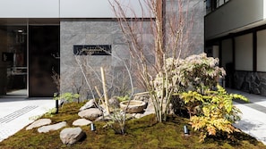 Un bâtiment moderne avec une façade en pierre, une enseigne indiquant «Hôtel Rhassare», et un jardin aménagé avec des rochers, de la mousse et des arbres.