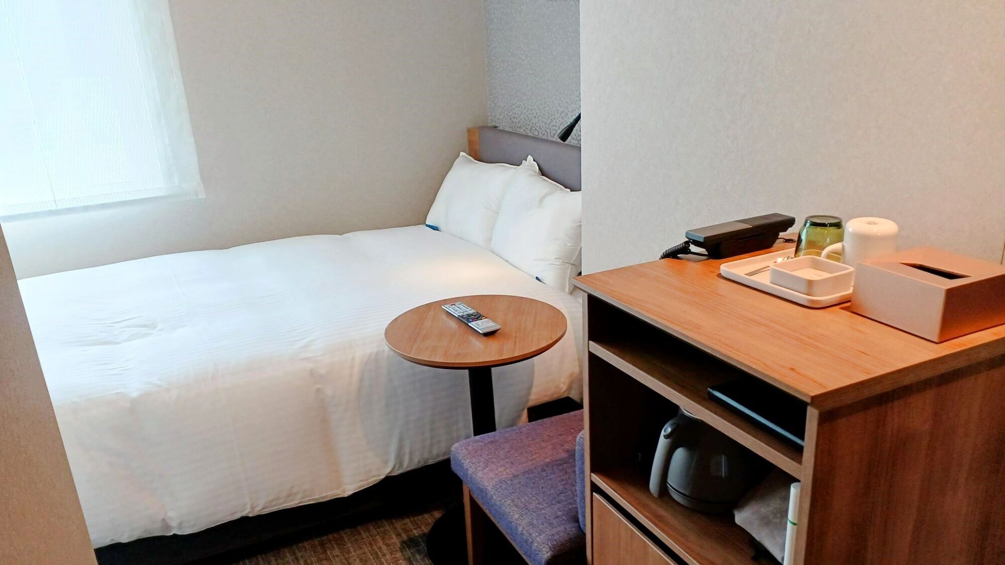 A hotel room with a bed, a small round table, a chair, a desk with a telephone, a coffee maker, and a waste bin.