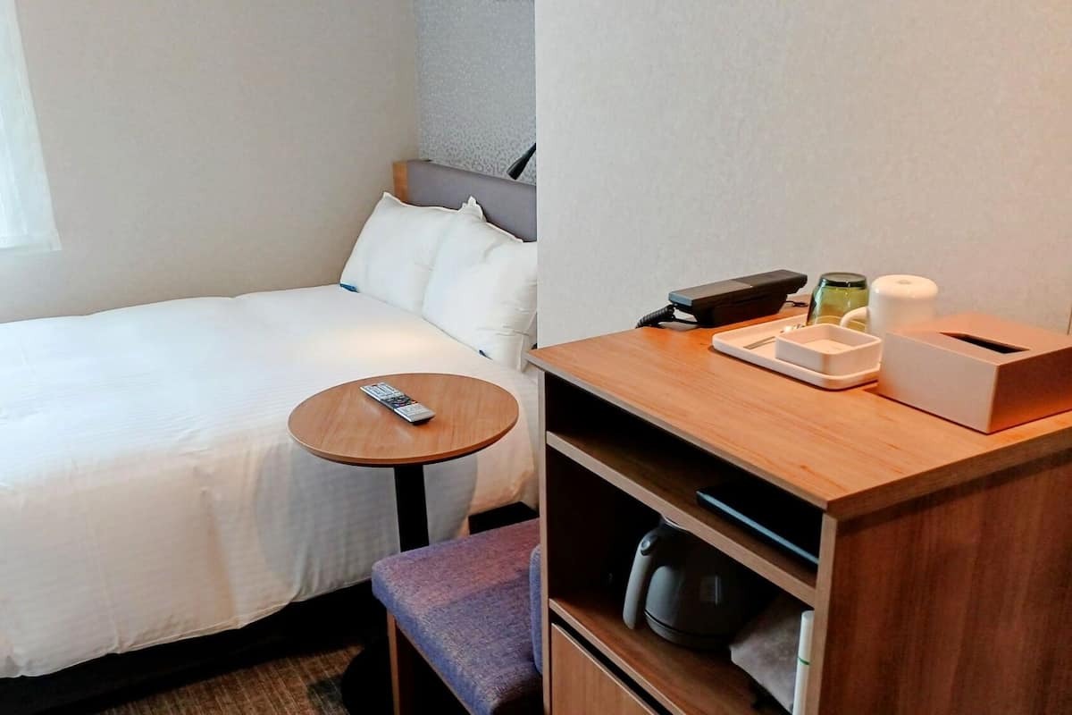 A hotel room with a bed, a small round table, a chair, a desk with a telephone, a coffee maker, and a waste bin.