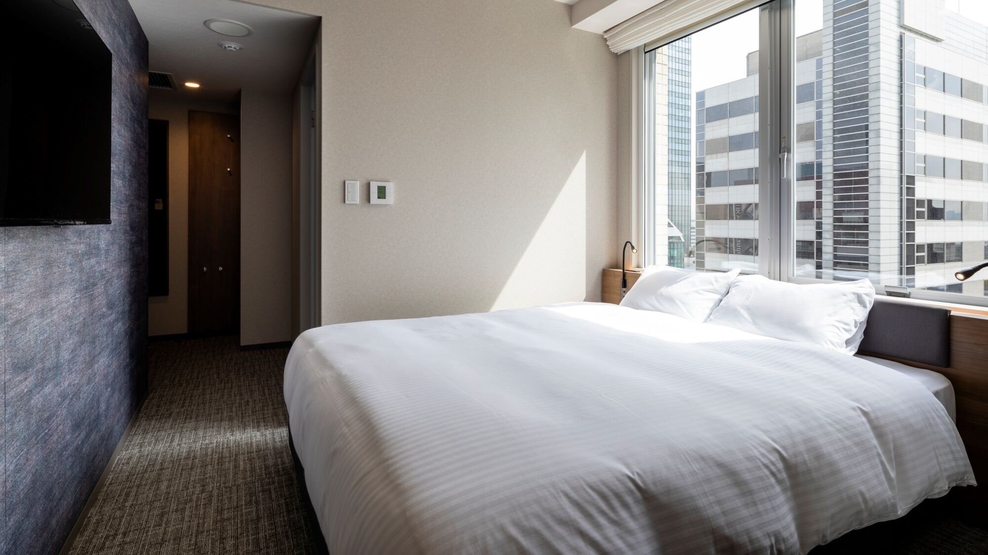 A hotel room with a large bed, a window with a city view, a bedside table with a lamp, and a door leading to another room.