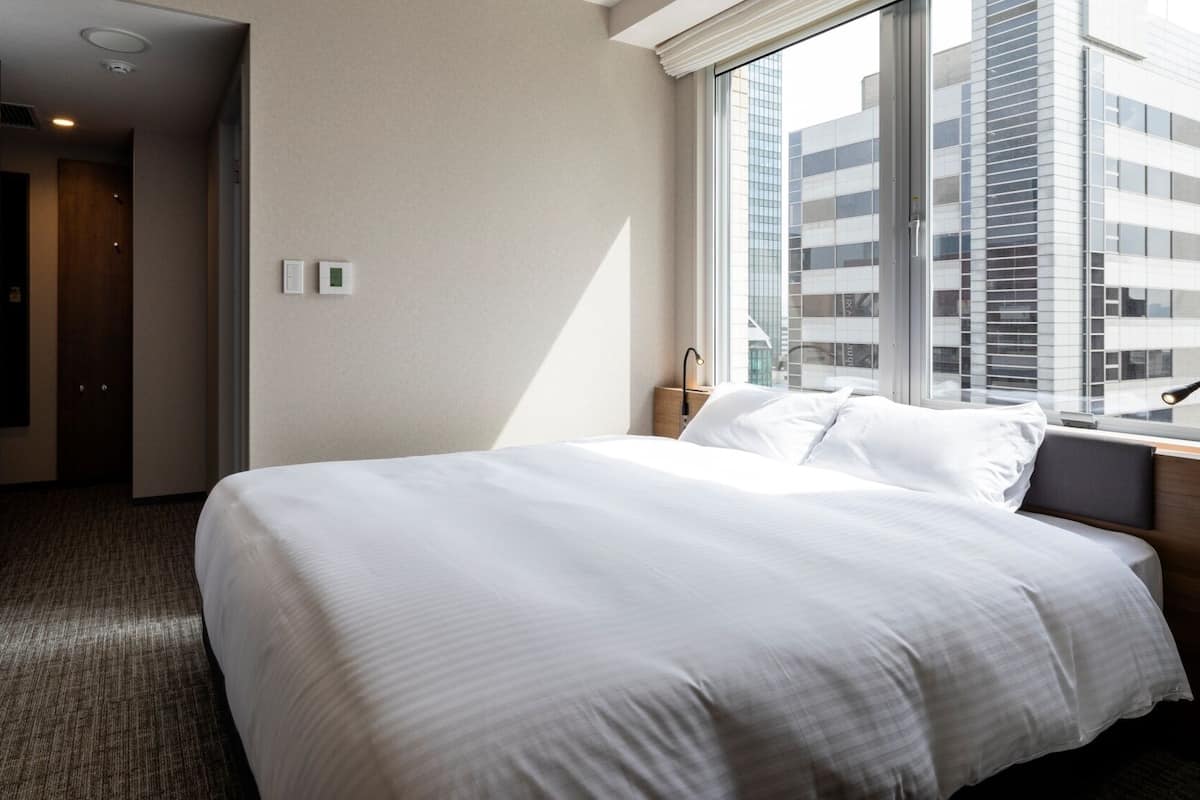 A hotel room with a large bed, a window with a city view, a bedside table with a lamp, and a door leading to another room.