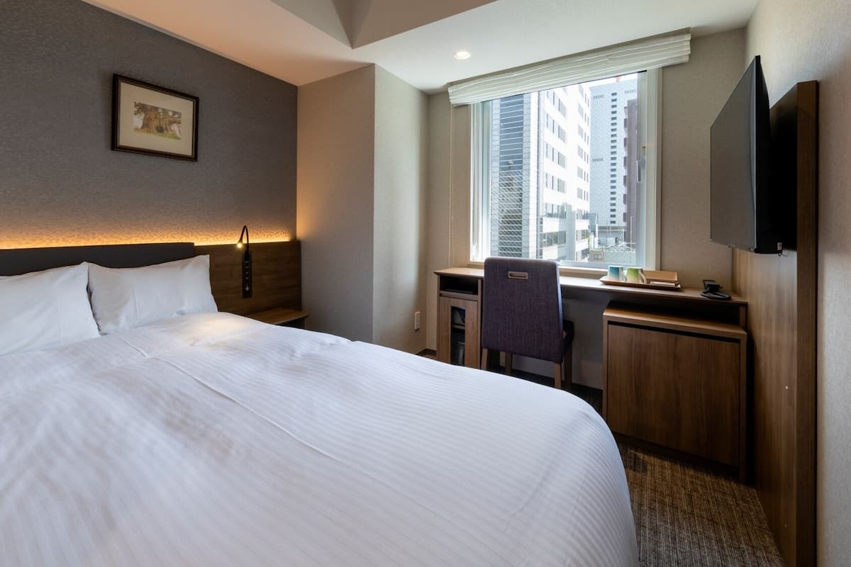 A hotel room with a large bed, a desk, a chair, a TV, and a window with a city view.