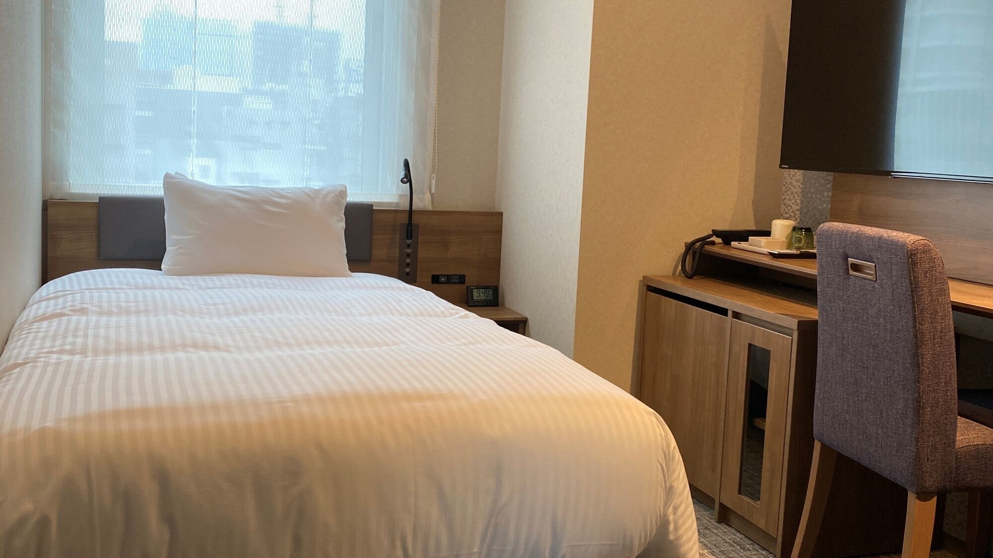 A hotel room with a bed, a desk, a chair, a TV, and a clock.