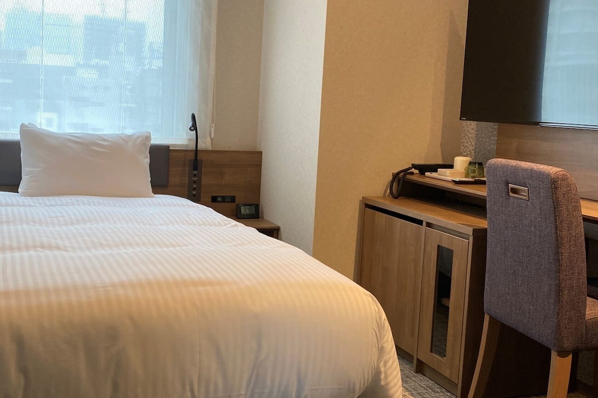 A hotel room with a bed, a desk, a chair, a TV, and a clock.