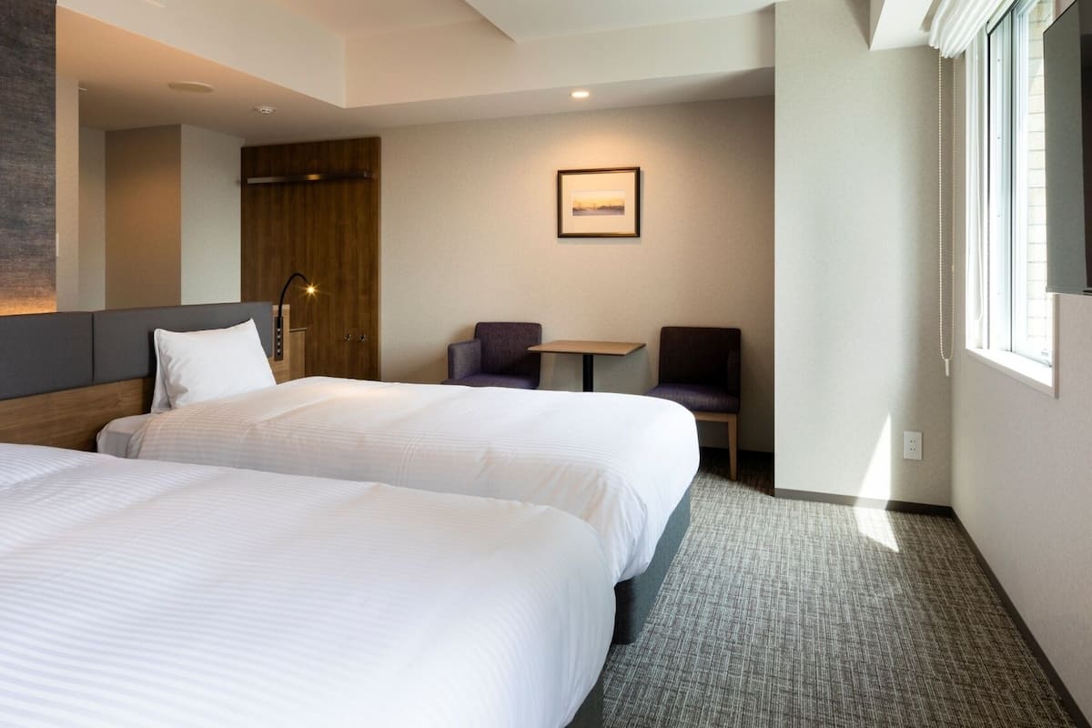 A hotel room with two beds, a desk, a chair, a framed picture on the wall, and a window.