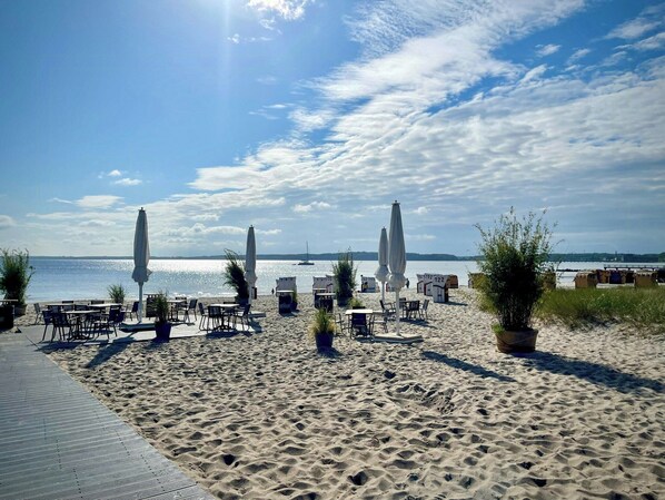 Ein Strandbereich mit Sitzgelegenheiten im Freien, Sonnenschirmen und Blick auf das Meer.
