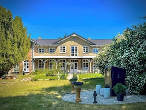 Ein großes, zweistöckiges Haus mit einer symmetrischen Fassade, einer Veranda und einem gepflegten Garten mit unterschiedlichen Pflanzen und einer Statue.