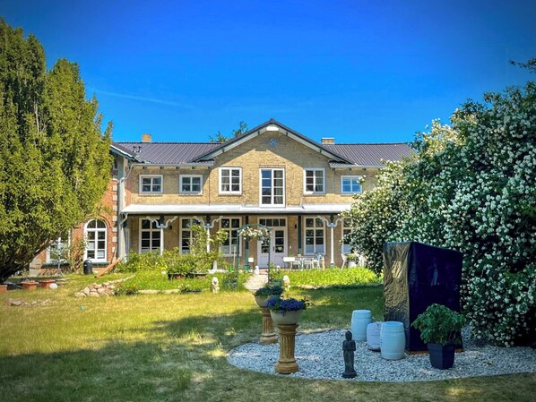 A large, two-story house with a symmetrical facade, a front porch, and a well-maintained garden with various plants and a statue. - vacation apartment with artistic charm (Prasdorf)
