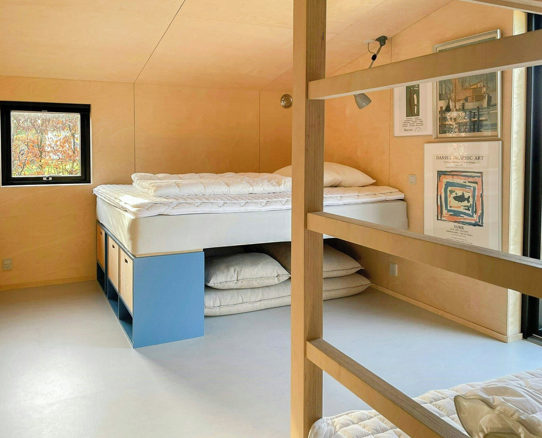 Quarto com beliche, uma moldura com arte gráfica dinamarquesa, uma janela com vista para árvores e um armário azul.