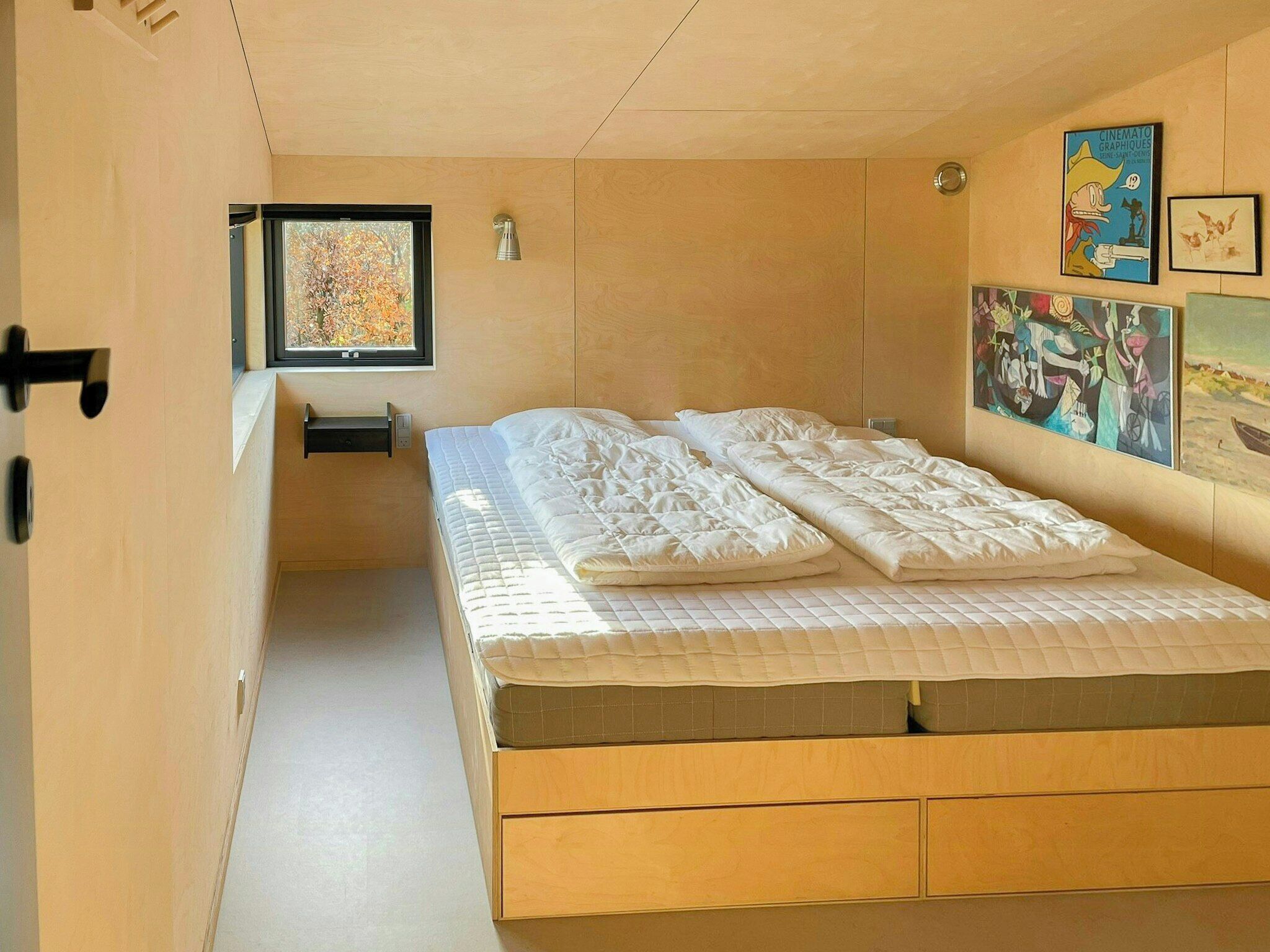 A bedroom with a bed, a window, a wall-mounted light, and artwork.