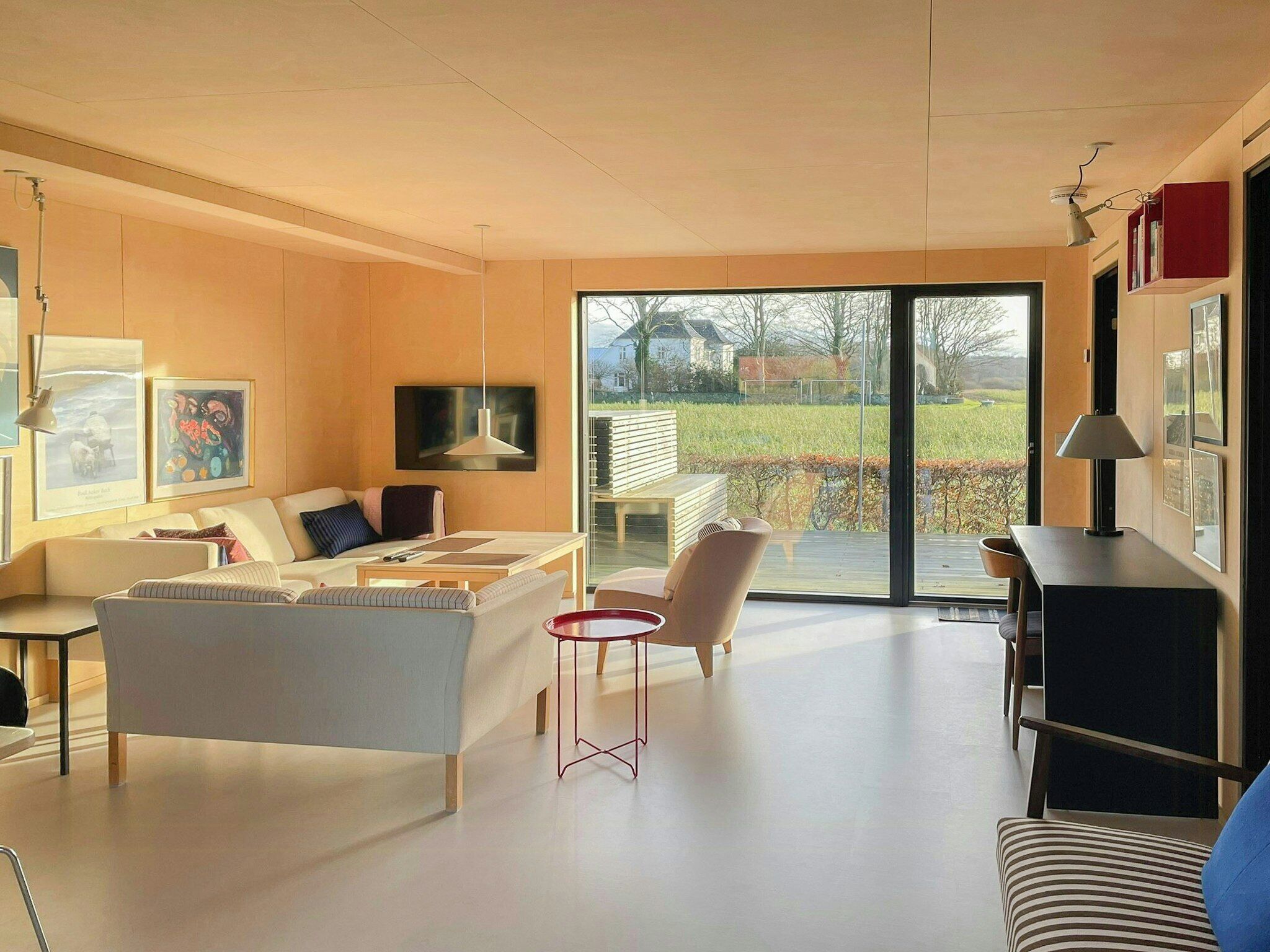 Un salon moderne avec un canapé, un fauteuil et une petite table. Une grande fenêtre offre une vue sur un jardin et une maison.