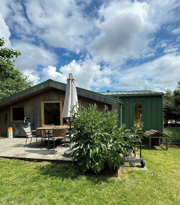 A wooden cabin with a patio, a grill, and a green shed.